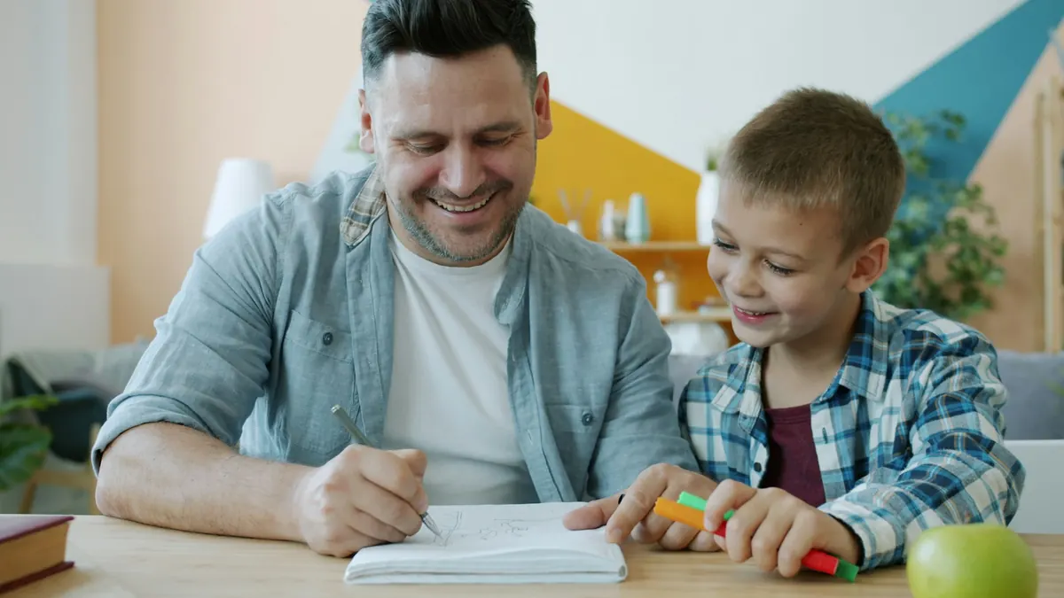 A smiling man and a young boy working on schoolwork together at a table