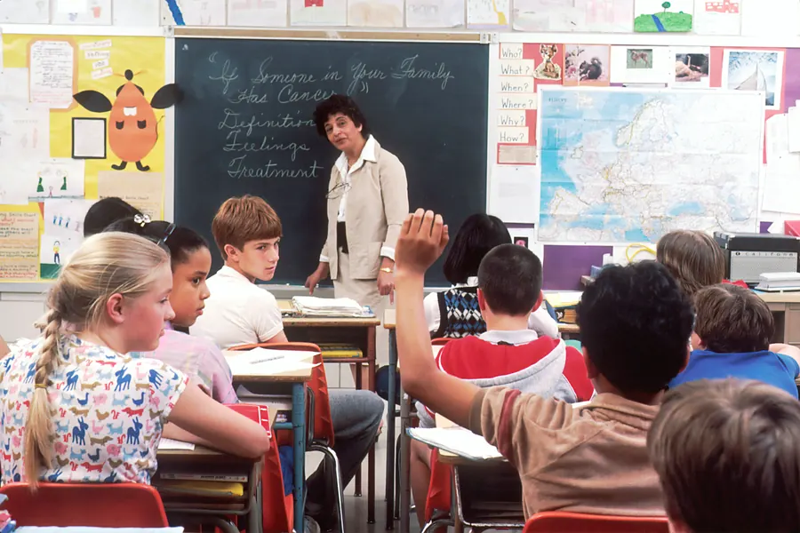 Students raising their hands in a classroom while their teacher stands at the chalkboard