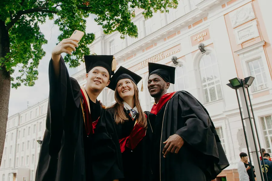 Three graduates in caps and gowns taking a selfie outside a university